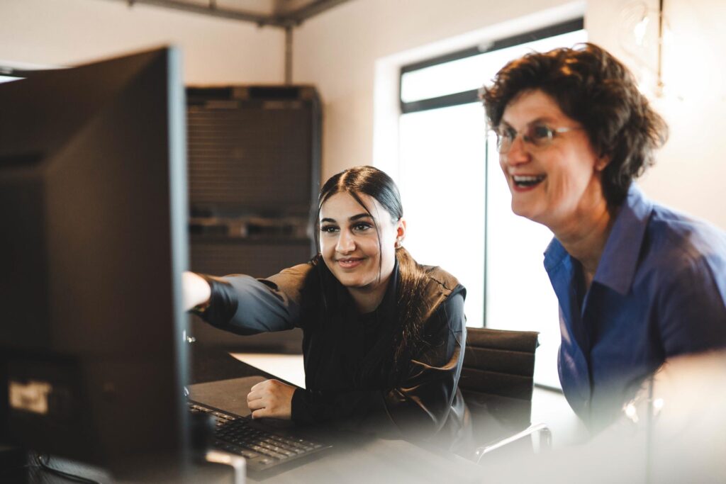 Zwei Mitarbeitende von STROHBECKE arbeiten gemeinsam am Computer und lachen – Symbol für Teamgeist, Wissenstransfer und Freude an der Arbeit.