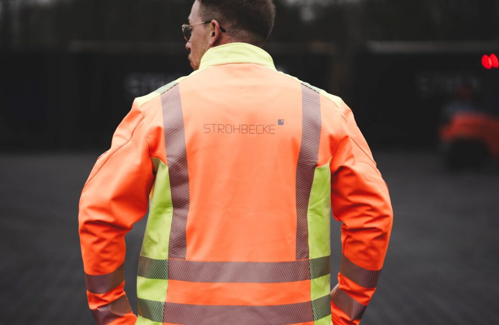 Eine Person trägt eine orange-gelbe Sicherheitsjacke mit dem STROHBECKE-Logo auf dem Rücken – Symbol für Einsatzbereitschaft und Verantwortung in der Logistik.